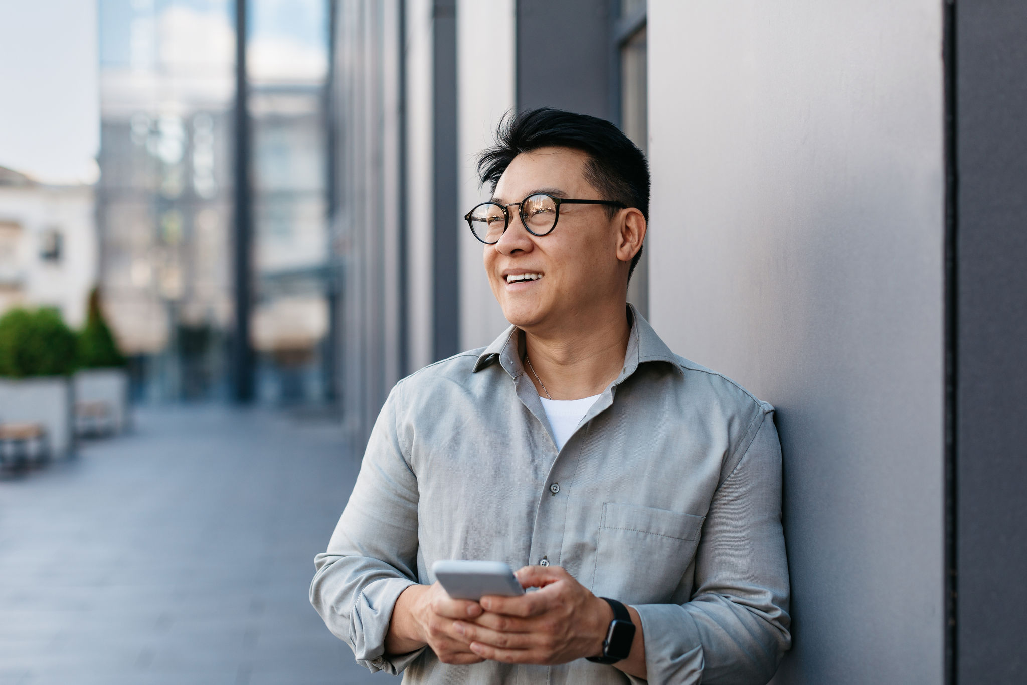 Confident korean middle aged businessman holding smartphone and looking aside at free space, standing near office building, outdoors. Male entrepreneur with gadget