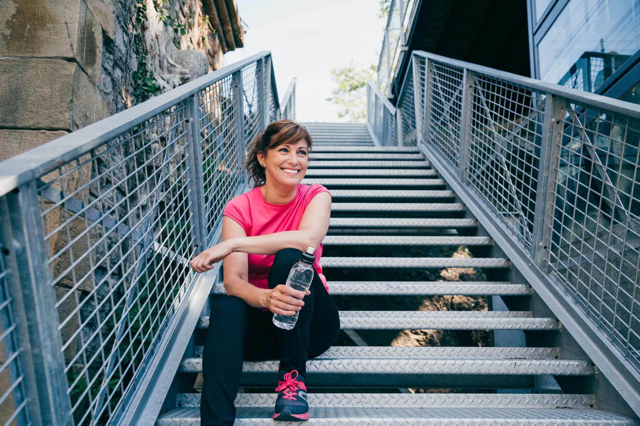 Happy and beautiful middle aged woman sitting on metallic stairs relaxing before running outdoors holding a water bottle