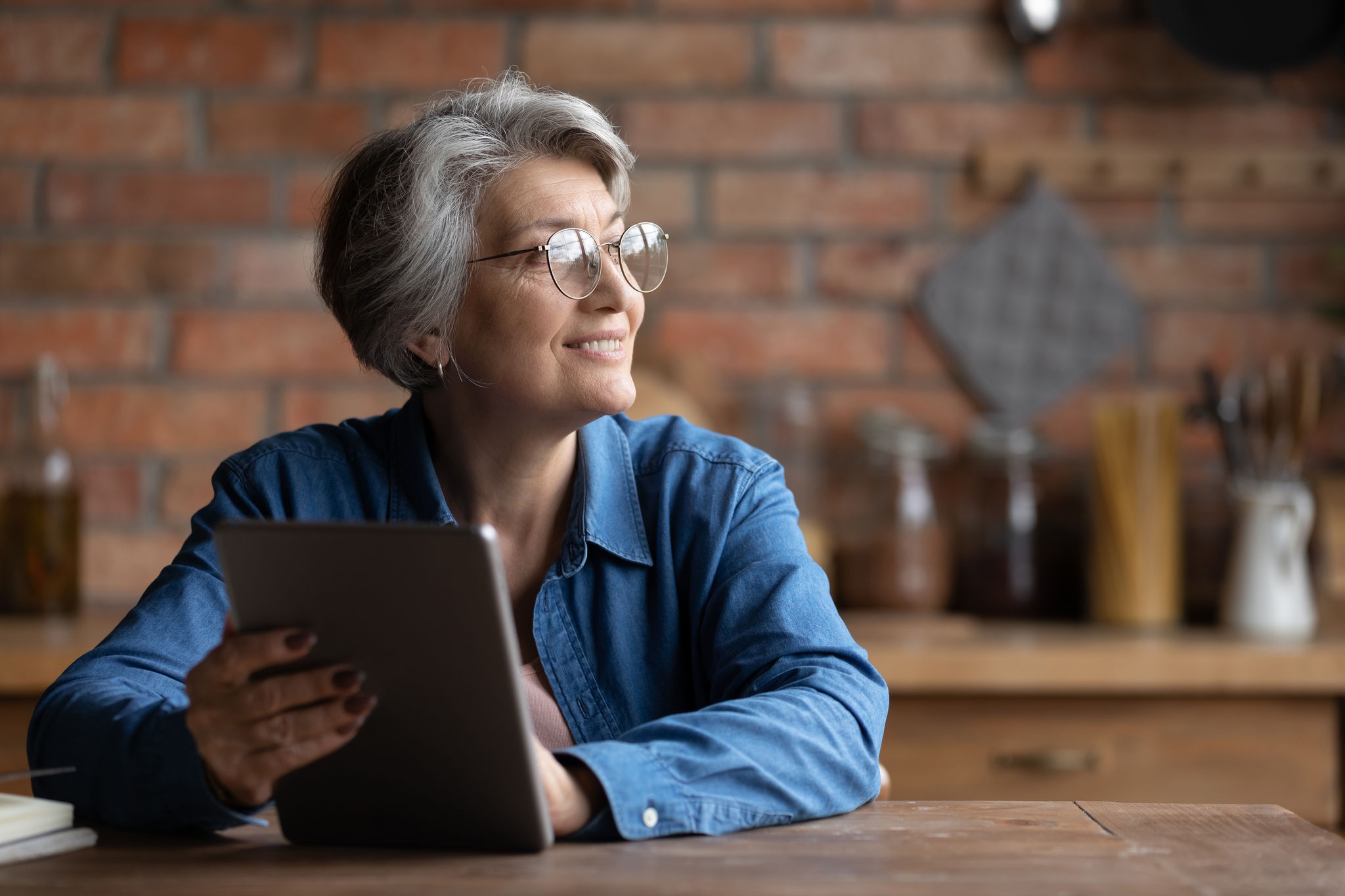 Dreamy senior grey haired woman in spectacles holding digital computer tablet in hands, looking in distance, copy space for advertising text of retired old people and modern technology easy usage.