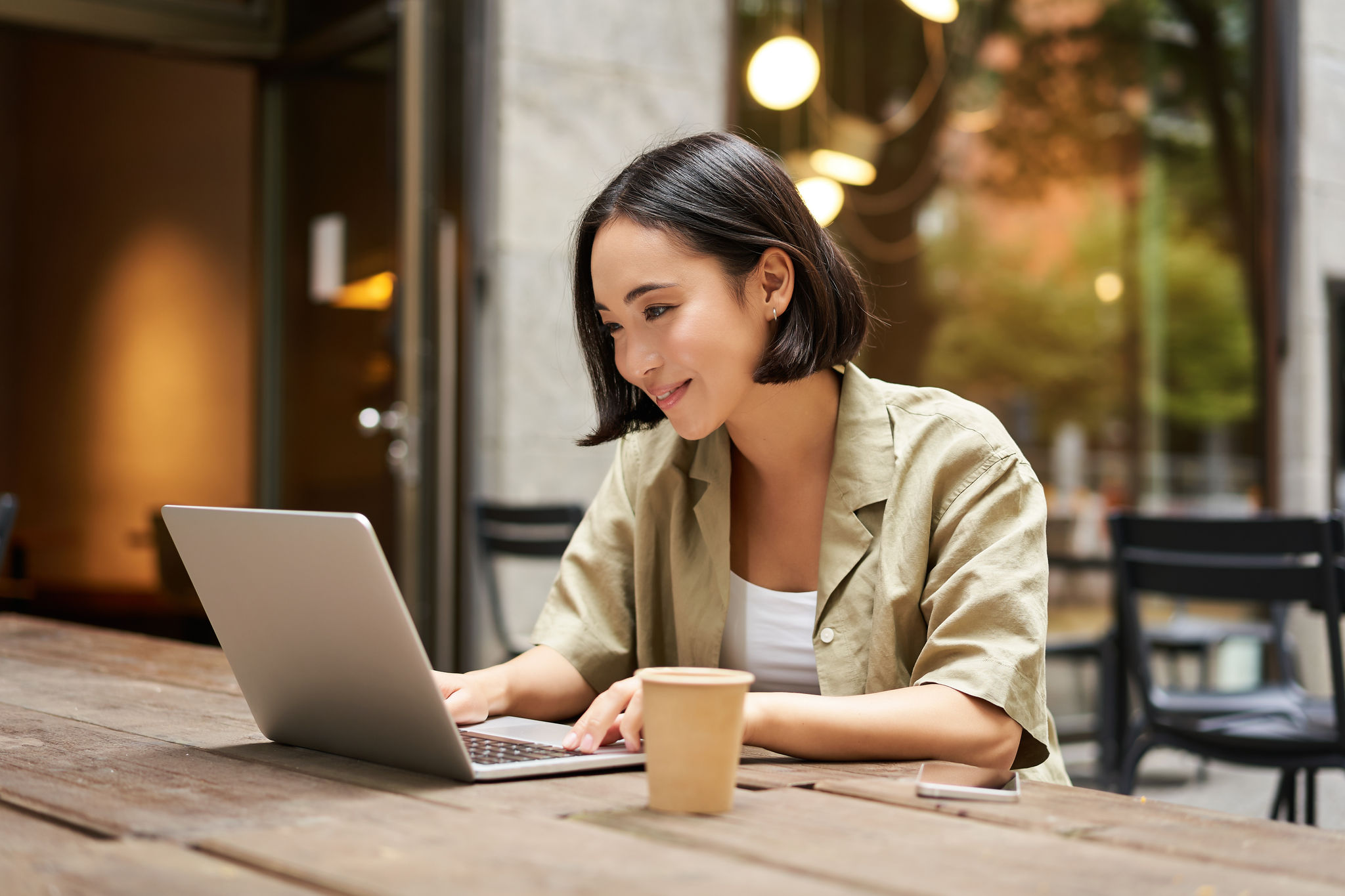 Young asian woman, digital nomad working remotely from a cafe, drinking coffee and using laptop, smiling.