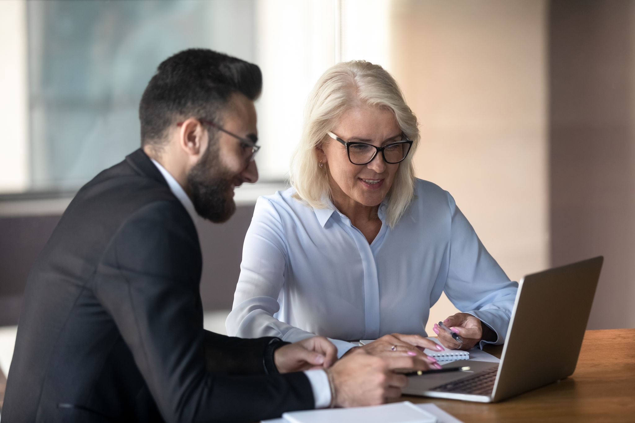 Mature businesswoman and young colleague working on project together, employees using laptop, discussing strategy, planning, senior mentor training teaching trainee, manager consulting client